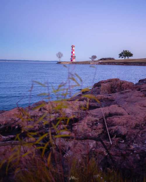A lighthouse stands on the shore, surrounded by calm water and rocky terrain, under a soft twilight sky.
