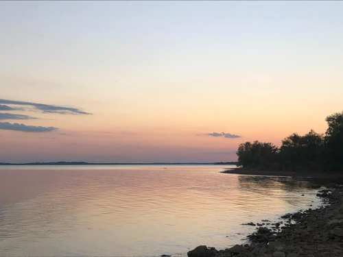 A serene lakeside view at sunset, with calm waters reflecting soft pastel colors and a tree-lined shore.
