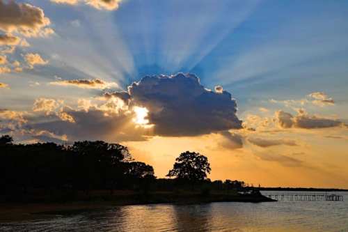 Sunset over a lake, with rays of light streaming through clouds and silhouettes of trees along the shore.