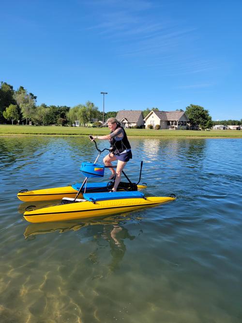 A person pedals on a yellow and blue watercraft on a calm lake, with houses and trees in the background.