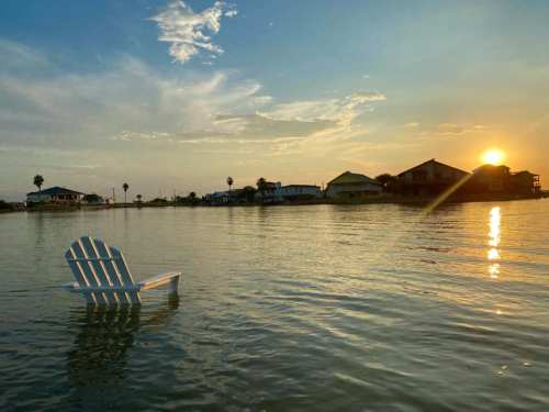 A white Adirondack chair floats on calm water at sunset, with houses and palm trees in the background.