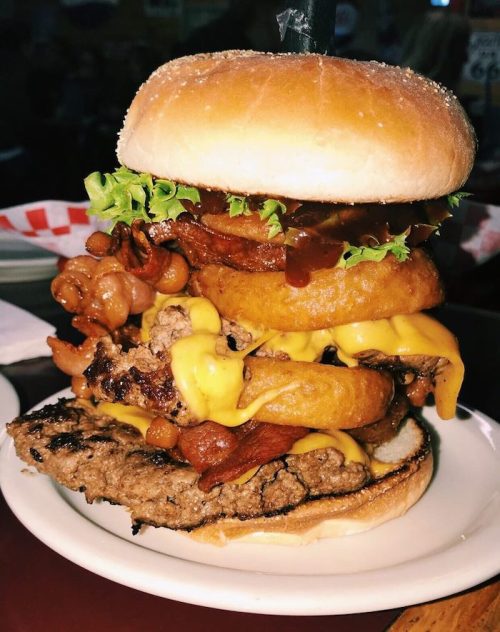 A towering burger with layers of beef, cheese, bacon, onion rings, lettuce, and barbecue sauce on a toasted bun.