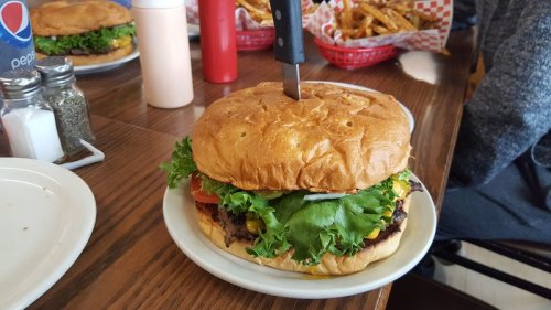 A large burger with lettuce, tomato, and cheese, topped with a knife, served on a plate with fries in the background.