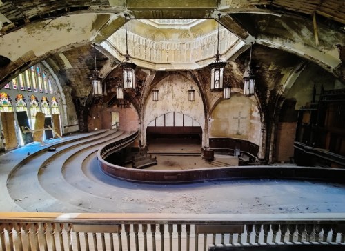 Abandoned church interior with curved seating, stained glass windows, and peeling walls, illuminated by hanging lights.