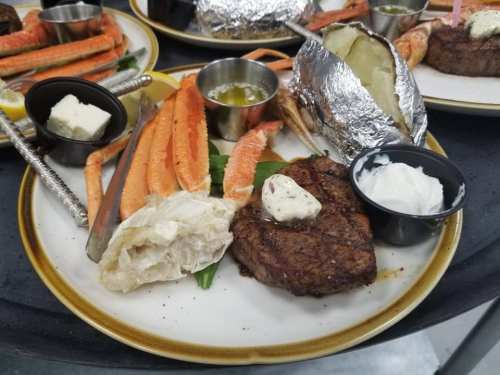 A plate featuring a steak with butter, crab legs, a baked potato, and dipping sauces, garnished with lemon.