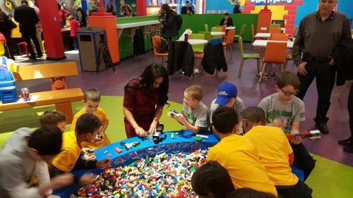A group of children and an adult play with colorful LEGO bricks in a vibrant, playful environment.