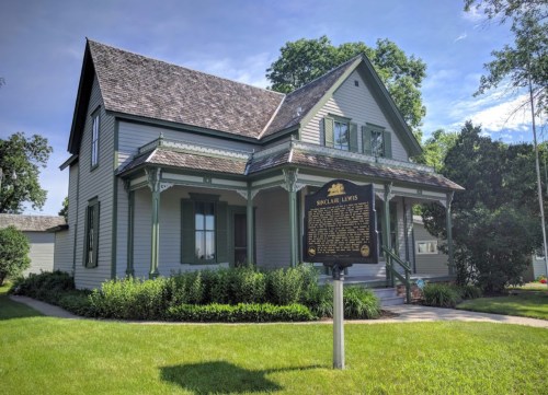 Historic house with green trim and a plaque in front, surrounded by lush grass and trees under a clear blue sky.