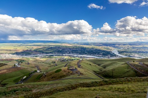 A panoramic view of rolling green hills, a river, and a small town under a partly cloudy sky.