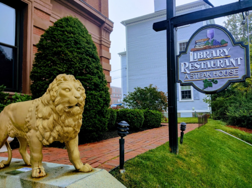 A golden lion statue stands near a sign for the Library Restaurant, a steakhouse, surrounded by greenery.