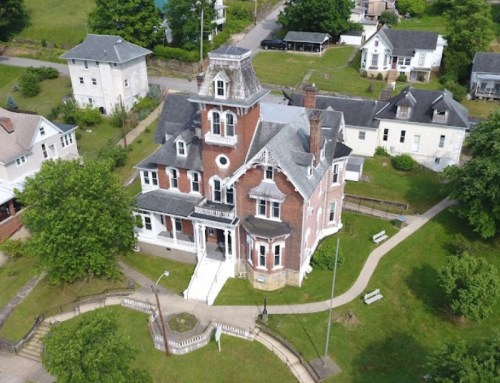 Aerial view of a large, historic brick house surrounded by green lawns and other buildings in a residential area.