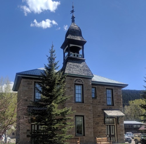Historic stone building with a clock tower, surrounded by trees and a clear blue sky.