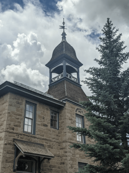 Historic stone building with a tall, ornate tower and a nearby evergreen tree under a cloudy sky.