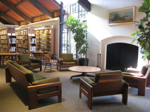 Cozy library lounge with wooden furniture, a round table, plants, and a fireplace, surrounded by bookshelves.