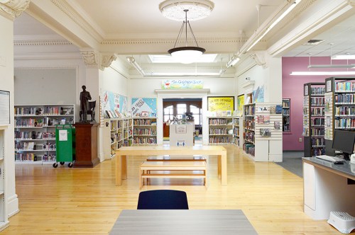 A spacious library interior with bookshelves, a central table, and a welcoming entrance.