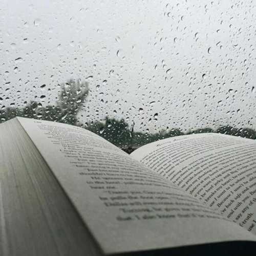 An open book rests on a surface, with raindrops on a window in the background, creating a cozy reading atmosphere.