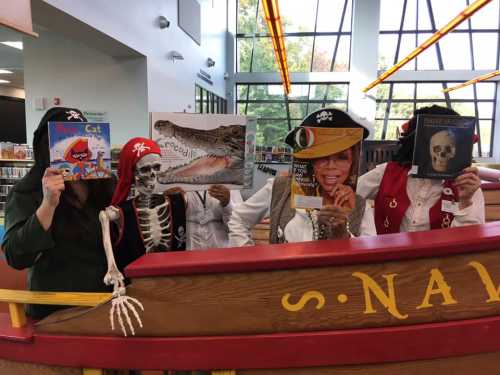 A group of people in pirate costumes holding books and a skeleton, posing in a library with a playful theme.