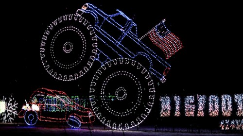 A festive display of a monster truck with oversized wheels, illuminated with colorful lights, against a dark background.