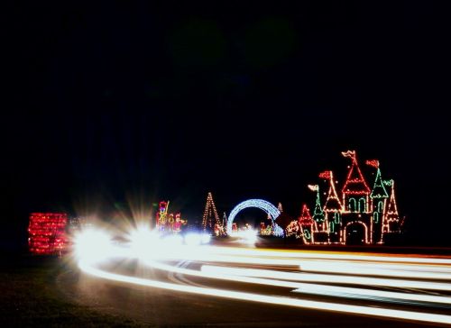 Colorful holiday lights illuminate a dark night, featuring festive shapes like a castle and trees, with car headlights in motion.