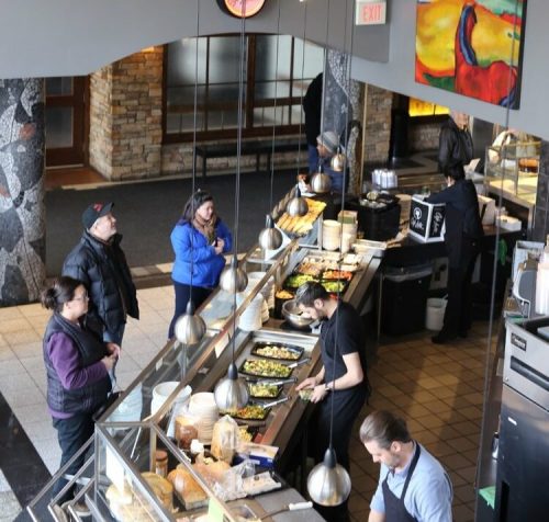 A bustling café interior with a long counter, staff preparing food, and customers enjoying the atmosphere.