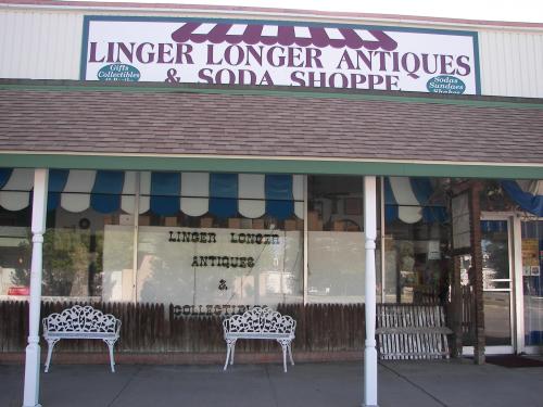 Exterior of Linger Longer Antiques & Soda Shoppe, featuring a sign and two white benches in front.