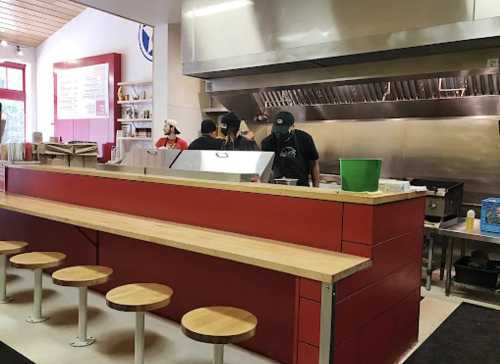 A modern restaurant interior with a red counter, wooden stools, and staff working behind a stainless steel kitchen.