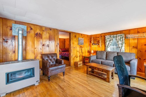 Cozy living room with wooden walls, a sofa, armchair, coffee table, and a wood stove, leading to a bedroom.