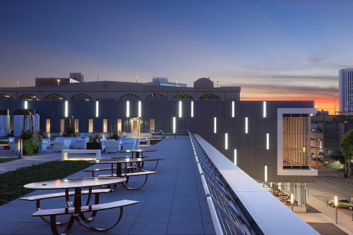 A rooftop terrace at dusk, featuring modern seating and city buildings in the background under a colorful sky.