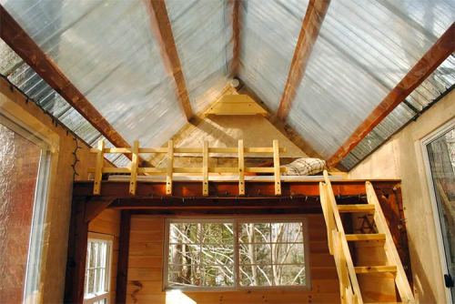 Interior of a cozy cabin with a loft bed, wooden ladder, and large windows, featuring a clear, sloped roof.