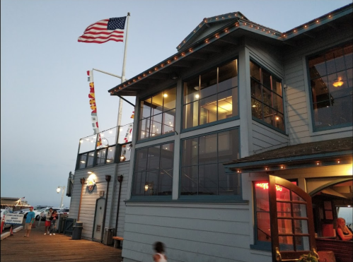 A waterfront building with large windows, an American flag, and colorful flags, illuminated at dusk.