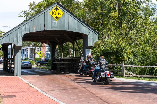 Motorcycles passing under a covered bridge with a height limit sign, surrounded by trees and a brick pathway.