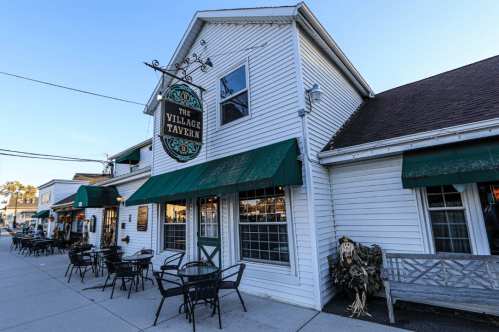 Exterior of The Village Tavern, featuring green awnings, outdoor seating, and a decorative scarecrow.