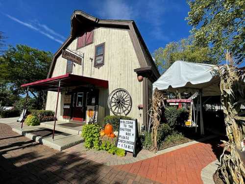 A rustic barn-style building with a welcome sign, pumpkins, and a tent outside, surrounded by trees and blue sky.