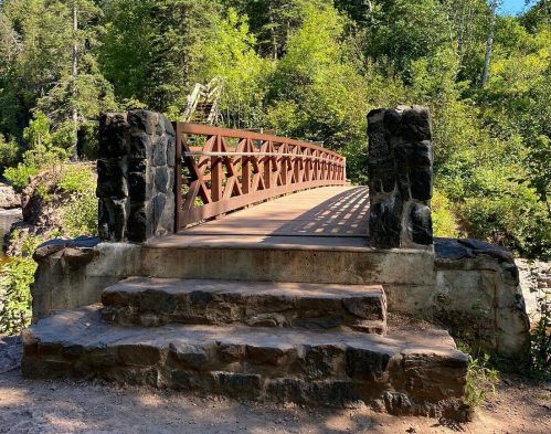 A wooden bridge with stone supports spans a lush, green forested area under a clear blue sky.