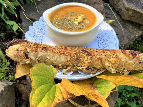 A bowl of soup garnished with herbs beside a crispy, golden-brown breadstick on a decorative plate.