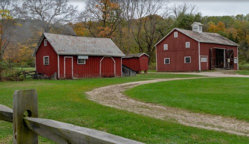 Two red barns sit beside a winding gravel path, surrounded by green grass and autumn trees in the background.