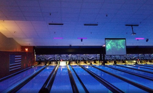 A dimly lit bowling alley with glowing lanes and a large screen displaying visuals. An American flag is visible on the wall.