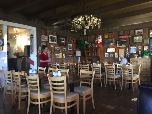 Cozy restaurant interior with wooden walls, tables, and vintage decor; a waitress serves while a customer sits at a table.