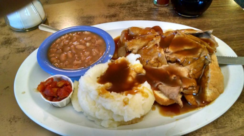 A plate of sliced roast beef with gravy, mashed potatoes, baked beans, and a side of diced tomatoes.