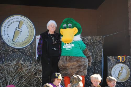 A woman stands next to a duck mascot in a green shirt at a duck calling contest event. Children are seated in front.