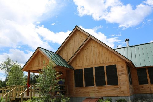 A wooden house with a green metal roof, surrounded by trees and under a blue sky with clouds.