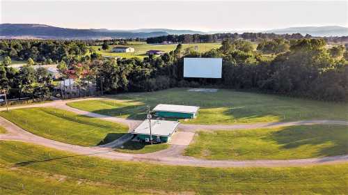 Aerial view of a drive-in movie theater with a large screen and grassy area, surrounded by trees and fields.