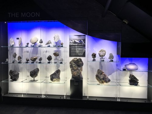 Display case featuring various lunar rocks and minerals, illuminated with blue lighting, labeled "The Moon."