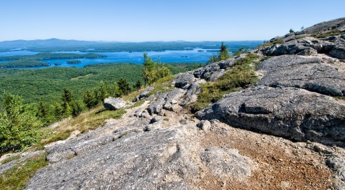 A panoramic view from a rocky mountain top overlooking a lush green landscape and a blue lake.