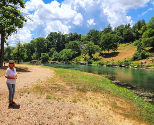 A woman stands by a riverbank surrounded by lush greenery and trees under a bright blue sky with fluffy clouds.