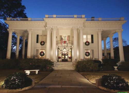 A grand white mansion with columns, decorated for the holidays, illuminated at dusk.