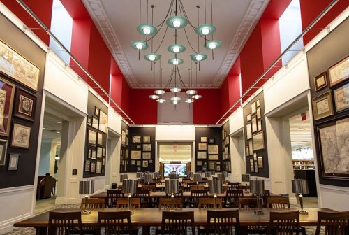 A spacious library interior with red walls, hanging lights, and tables surrounded by framed maps and artwork.