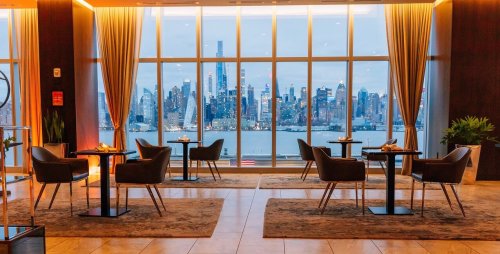 A stylish lounge with chairs and tables, featuring large windows showcasing a city skyline at dusk.