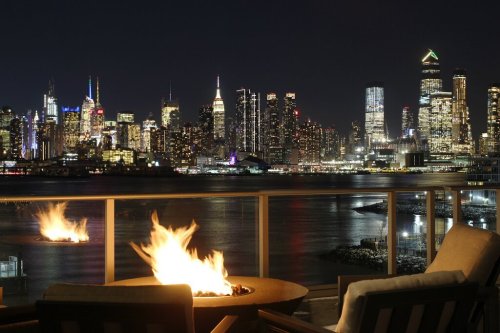A nighttime cityscape featuring a skyline with colorful lights, viewed from a balcony with a fire pit.