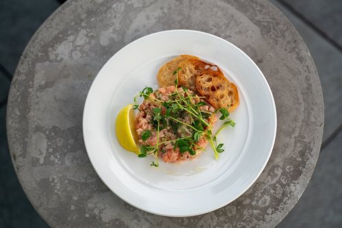 A plate of salmon tartare garnished with microgreens, served with lemon wedges and toasted bread on a stone table.