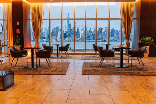 A modern lounge with chairs and tables, featuring large windows showcasing a city skyline at dusk.